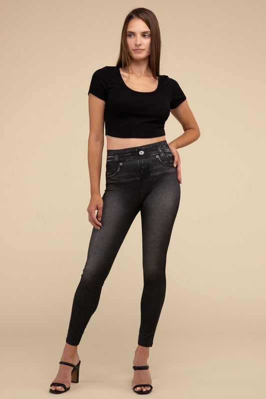 A person wearing Denim Leggings in a high-waisted black design and paired with black heels poses against a plain beige background, highlighting the sleek, stretchy fit.