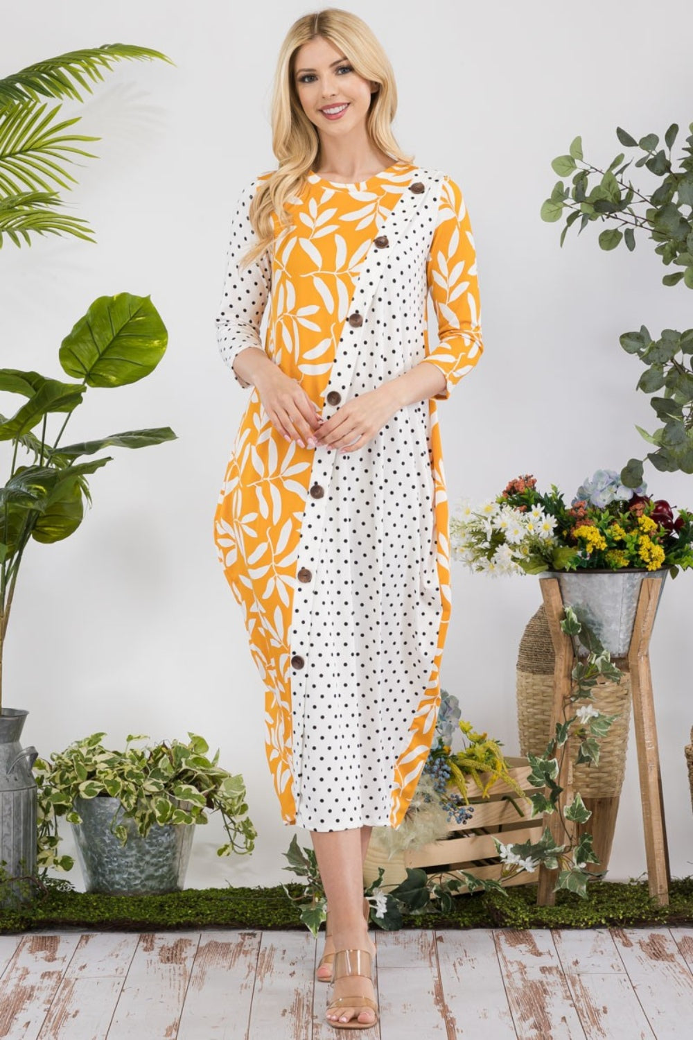 In a garden setting, a woman models the Celeste Full Size Floral Polka Dot Contrast Midi-Dress with Pockets, featuring its versatile yellow design among lush plants and vibrant blooms.