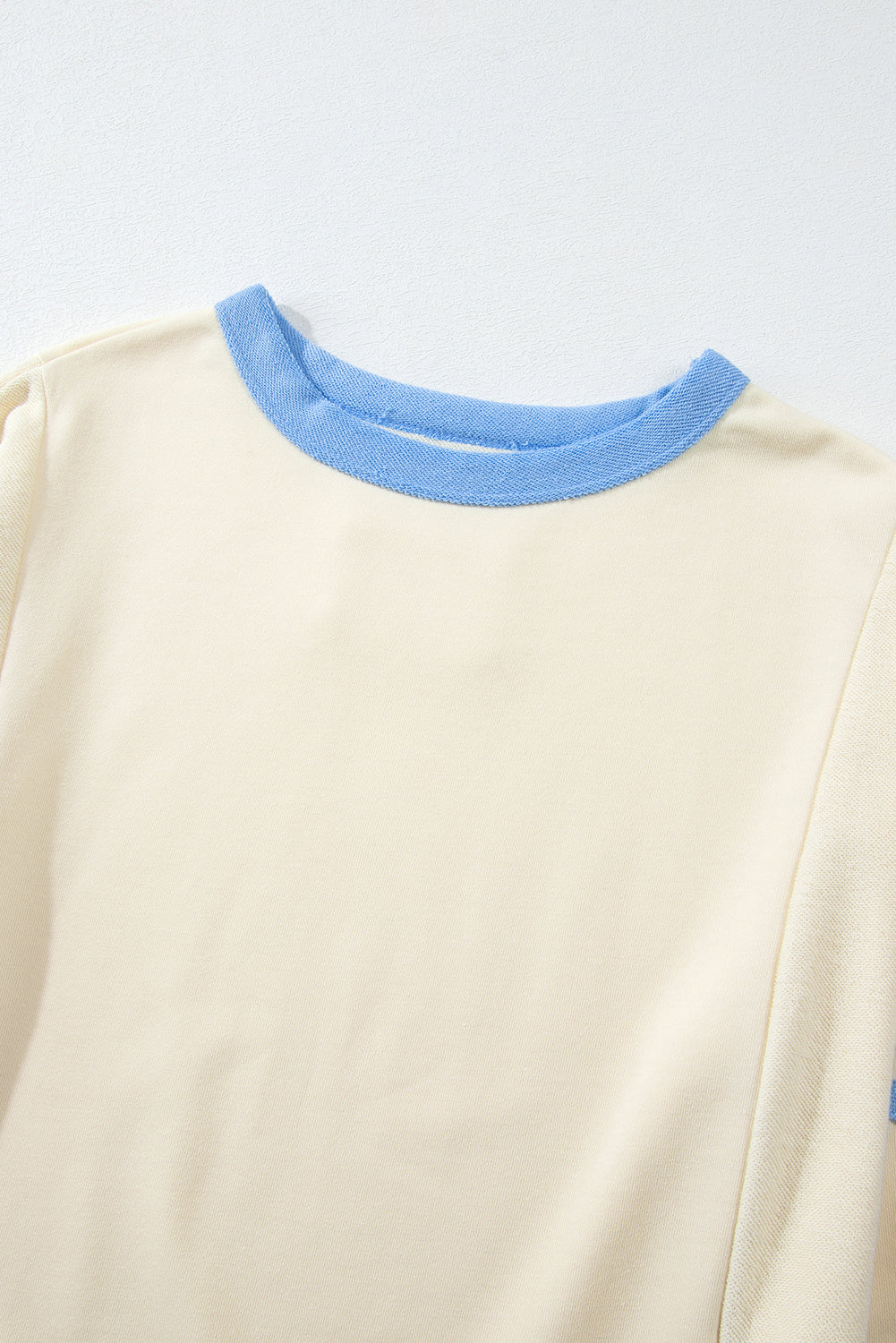 A person with long, wavy, light brown hair stands facing away, wearing the Beige Star Patchwork Exposed Seam Oversized Sweatshirt paired with denim shorts. The background is a plain, light peach wall, adding an edgy vibe to the look.