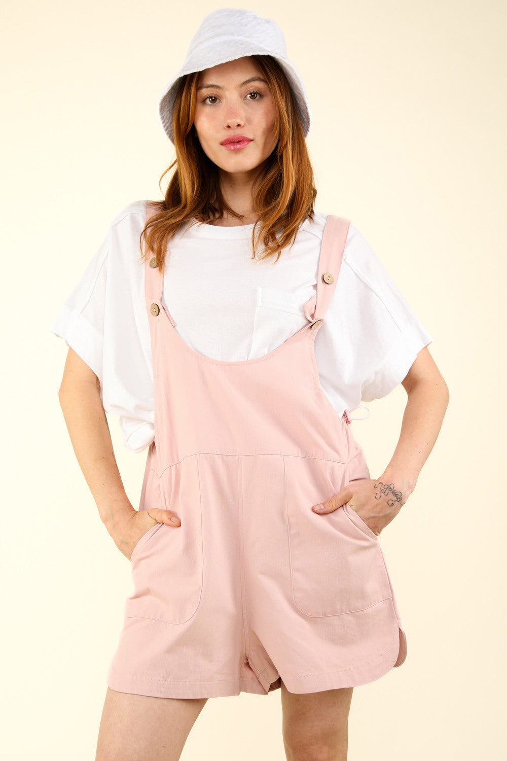 Person wearing a white hat, a white T-shirt, and VERY J Adjustable Waist Suspender Overalls with Pockets in light pink, poses with hands in the side pockets against a plain background.