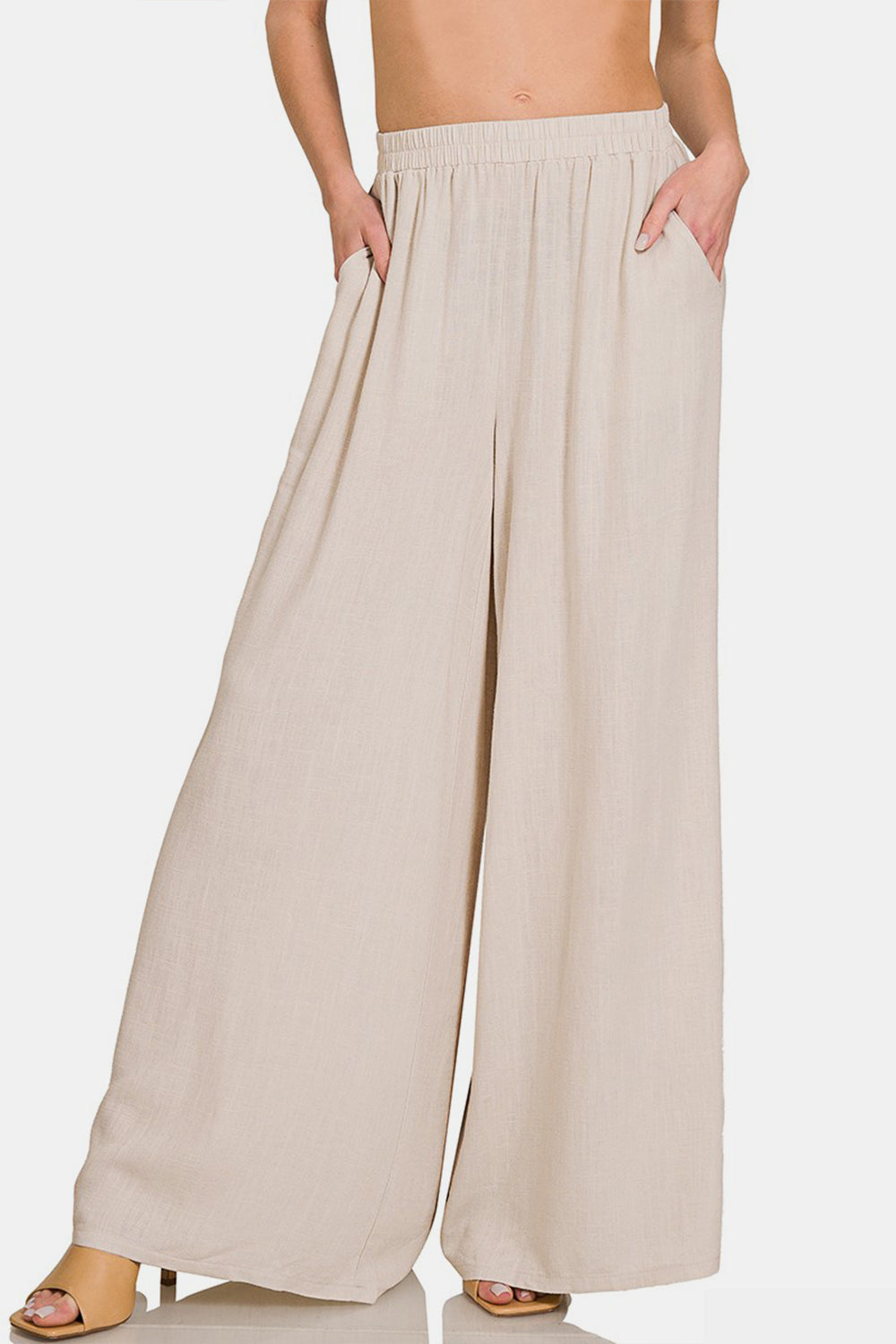 Person wearing Zenana Pleated Linen Blend Wide Leg Pants in beige with hands in pockets, shown from waist to feet. The pleated pants have an elastic waistband and flowy fabric. Model is also wearing light-colored open-toed sandals.