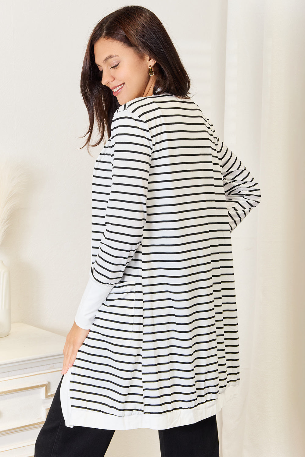 A woman wearing the Double Take Striped Open Front Longline Cardigan over a white top and black pants stands indoors, smiling at the camera. She is posing with one hand on her hip, showcasing this versatile piece.