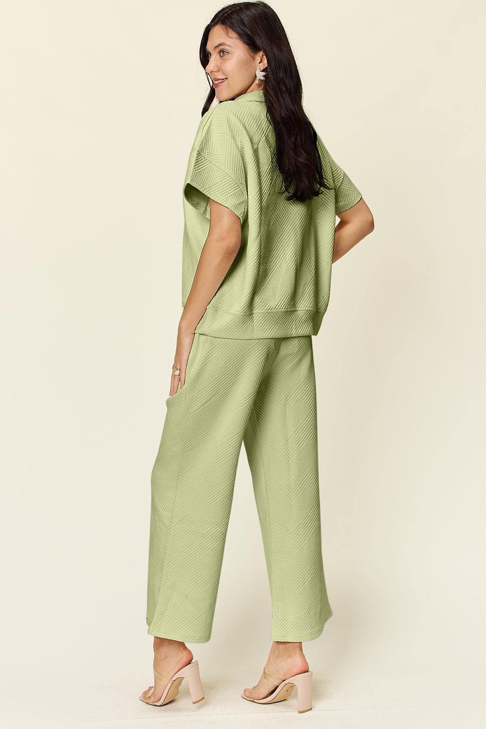 A woman is wearing the Double Take Full Size Texture Half Zip Short Sleeve Top and Pants Set, featuring a white zip-up short-sleeve top and matching wide-leg, drawstring pants. She stands with one hand in her pocket and smiles against a light background.