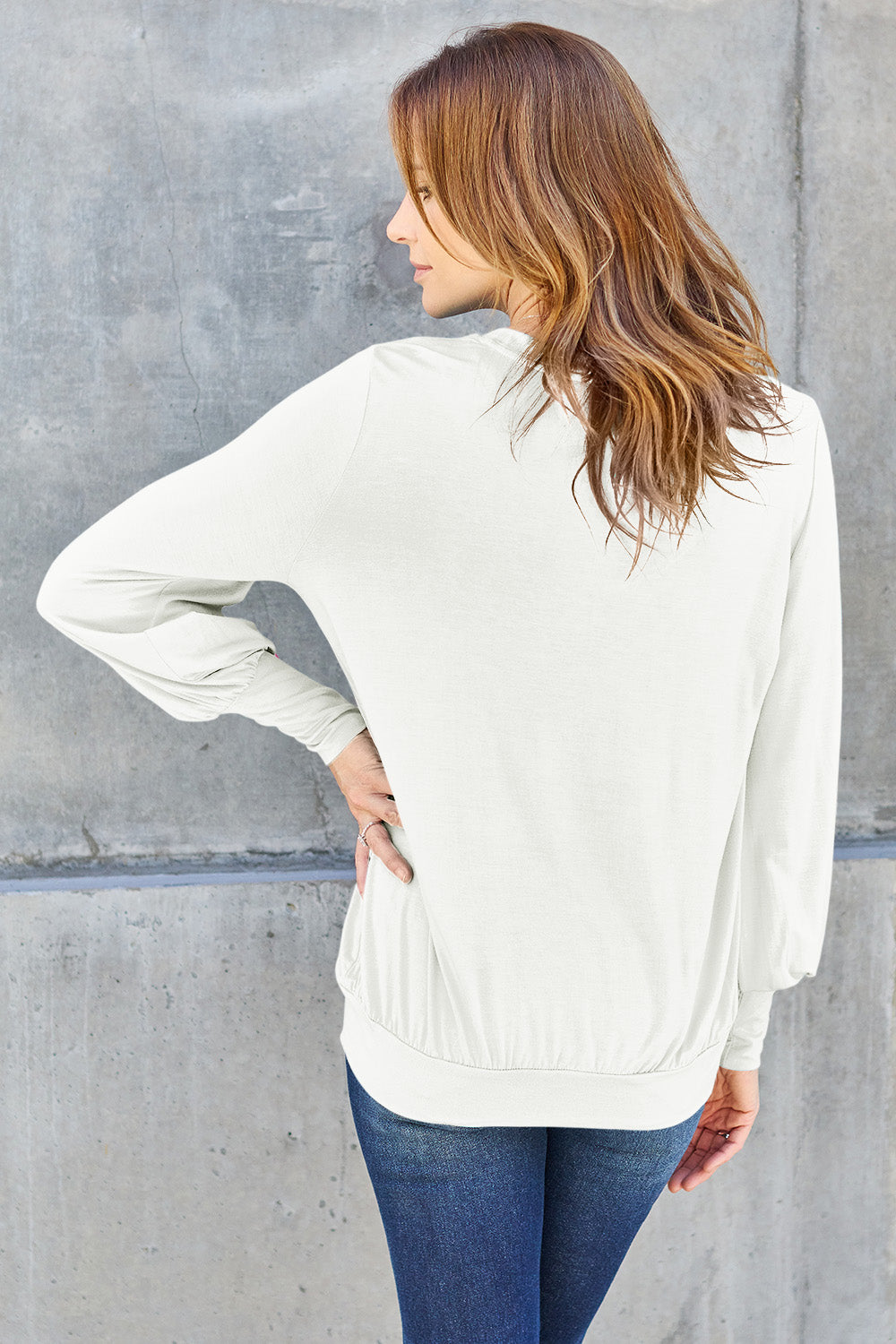 A woman with brown hair is standing against a concrete wall, wearing the Basic Bae Full Size V-Neck Lantern Sleeve Top in teal and blue basic-style jeans, smiling.