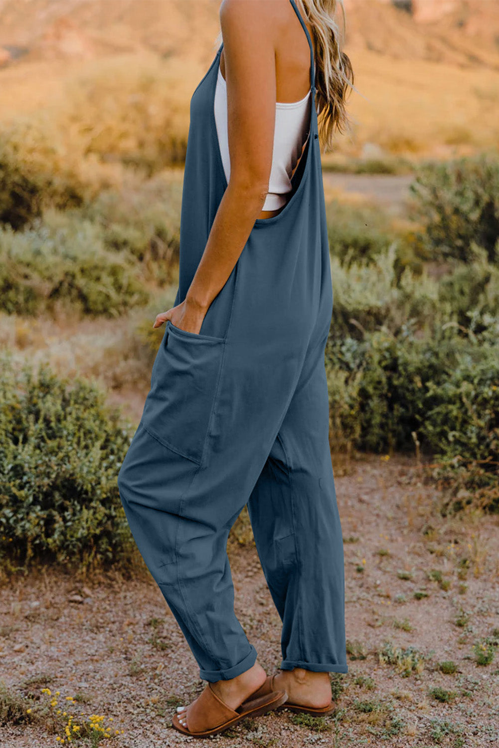 Person wearing a Double Take Full Size V-Neck Sleeveless Jumpsuit with Pockets and sandals, walking outdoors on a dirt trail with bushes in the background.