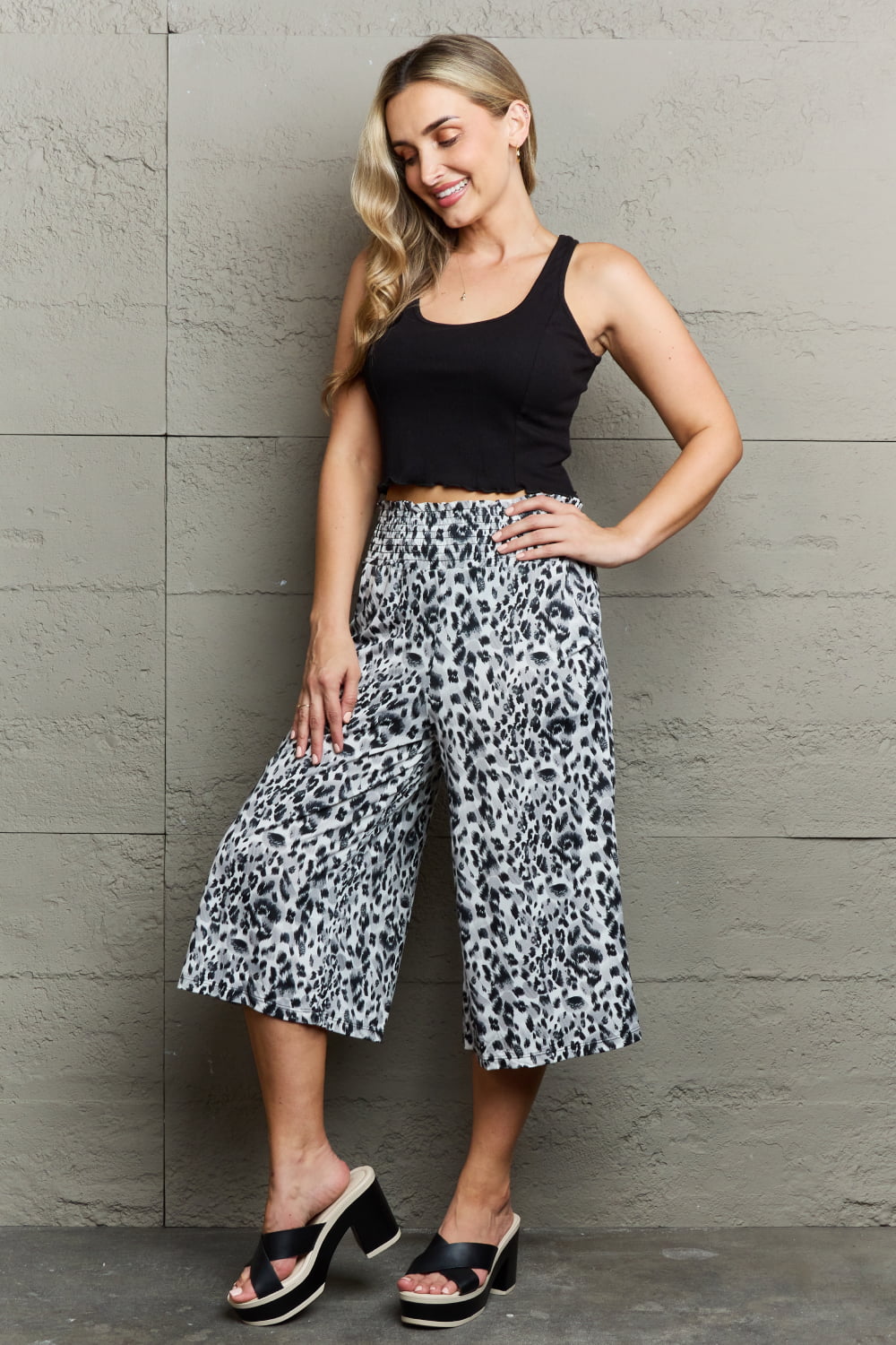 A person wearing Ninexis Leopard High Waist Flowy Wide Leg Pants with Pockets and black platform sandals, standing against a gray wall.