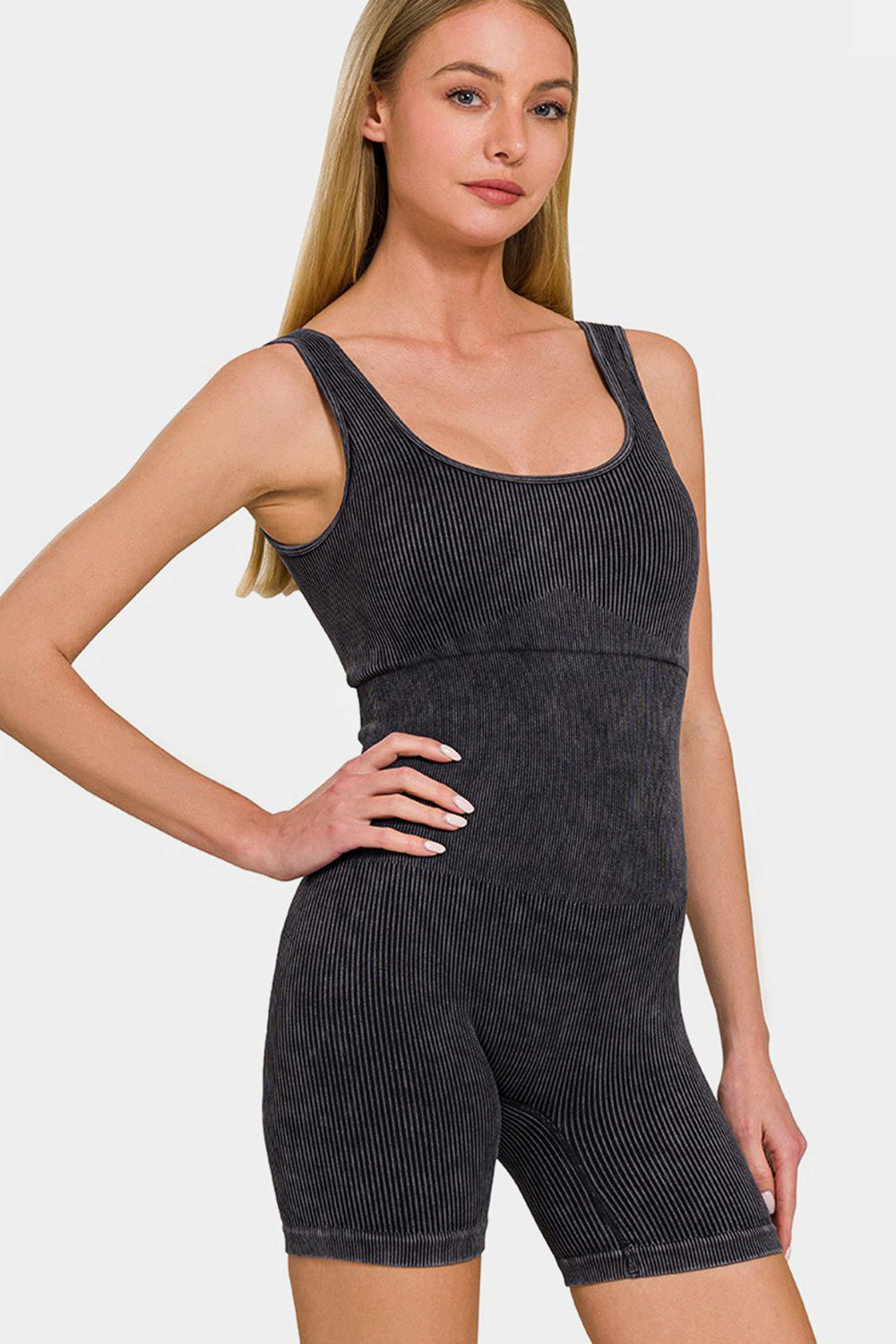 A person with long blonde hair sporting a versatile piece, the Zenana Washed Ribbed Romper with Pad, stands with one hand on their hip against a plain background.