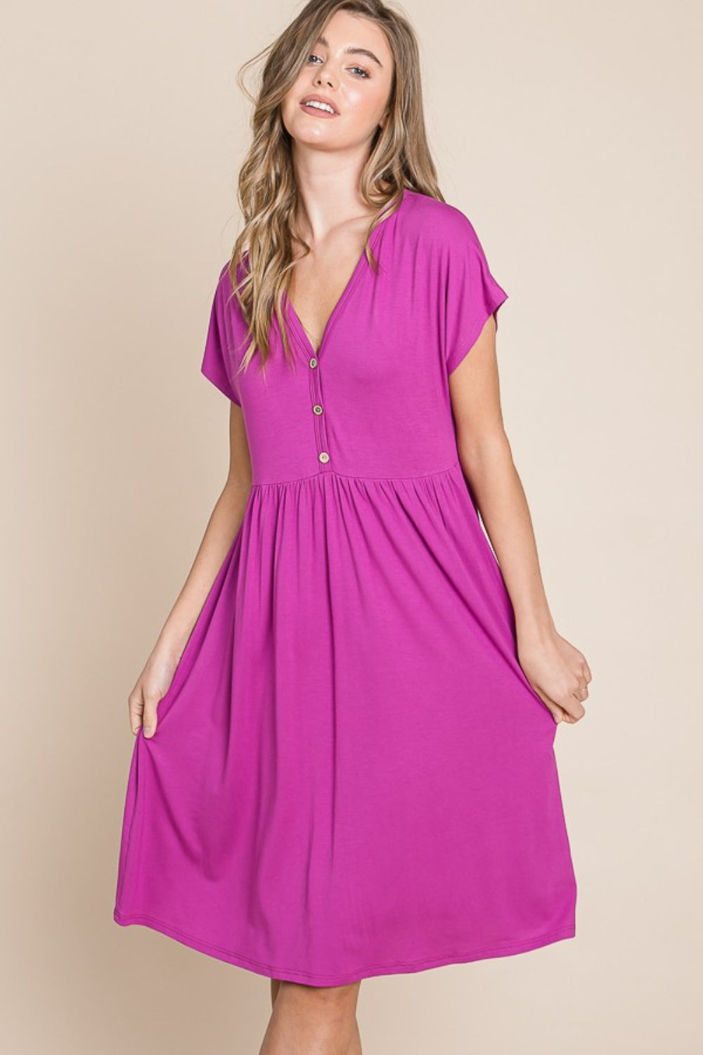 A person stands against a neutral background wearing the BOMBOM V-Neck Short Sleeve Dress, featuring a vibrant pink color and button details, crafted from soft rayon spandex fabric.