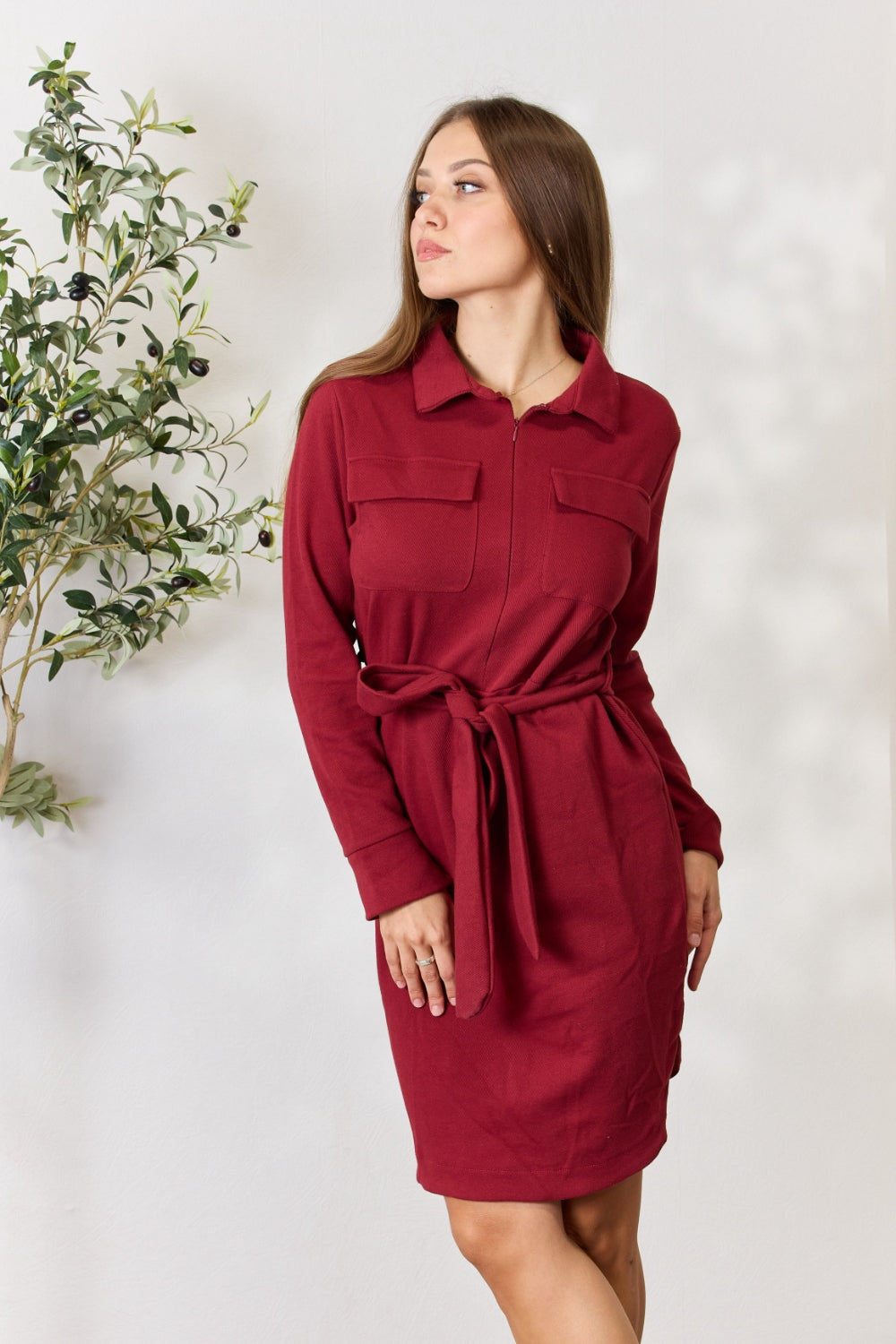 A woman wearing the Culture Code Full Size Tie Front Half Zip Long Sleeve Shirt Dress stands in front of a plain background next to a potted plant.