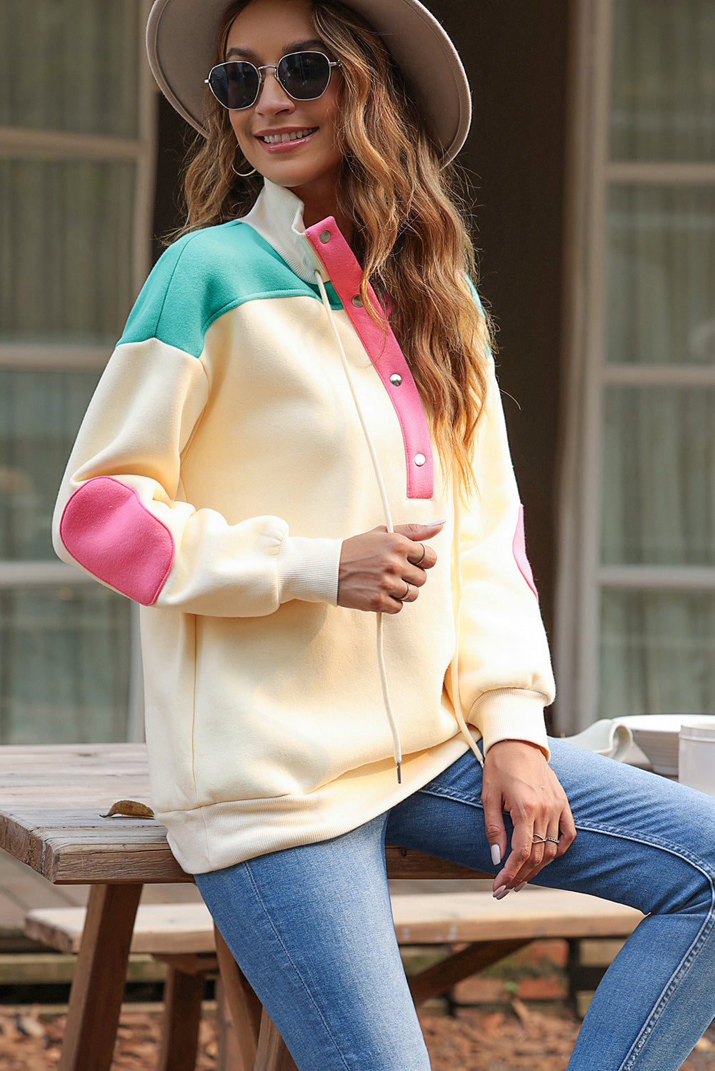 A woman wearing the Apricot Color Block Elbow Patch Half Button Sweatshirt, featuring cream and mint colors with pink elbow patches and a turn-down collar with snap button closure, stands outdoors. She has long wavy hair, sunglasses, a hat, and is wearing blue jeans.