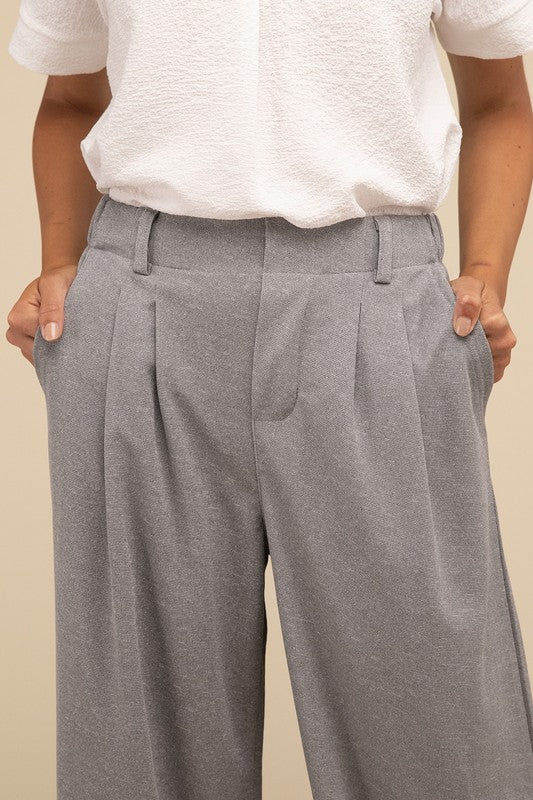 A woman stands against a beige background, highlighting her chic ensemble with a tailored fit. She wears a white short-sleeve shirt paired with versatile pink Waffle Trouser Pants, hands casually in pockets, complemented by stylish strappy sandals.