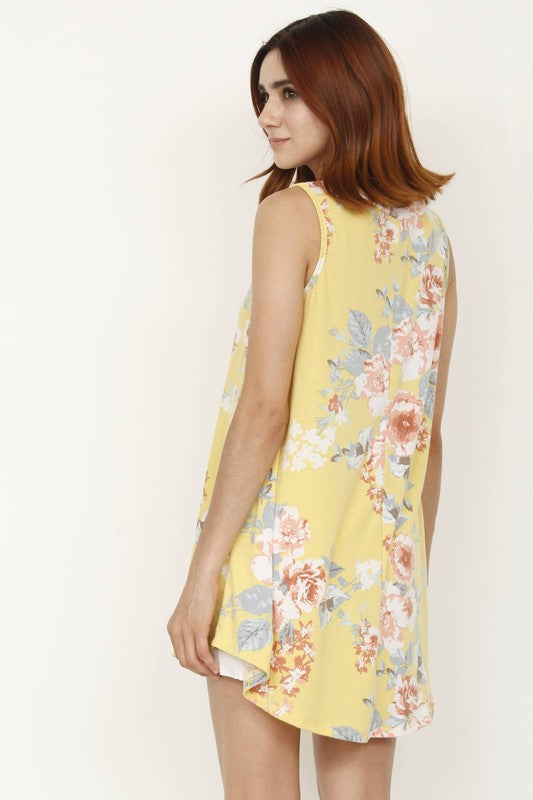 A woman wearing a Floral Tank Top with a yellow floral print stands outdoors near a white wall and plants.