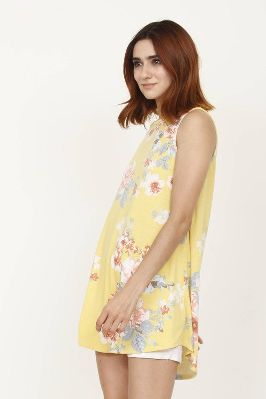 A woman wearing a Floral Tank Top with a yellow floral print stands outdoors near a white wall and plants.