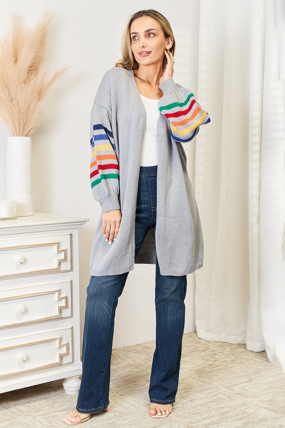 A person wearing the Angel Wings Multicolored Stripe Open Front Longline Cardigan over a white top and blue jeans stands indoors near a white cabinet, showcasing a fashionable cozy look.