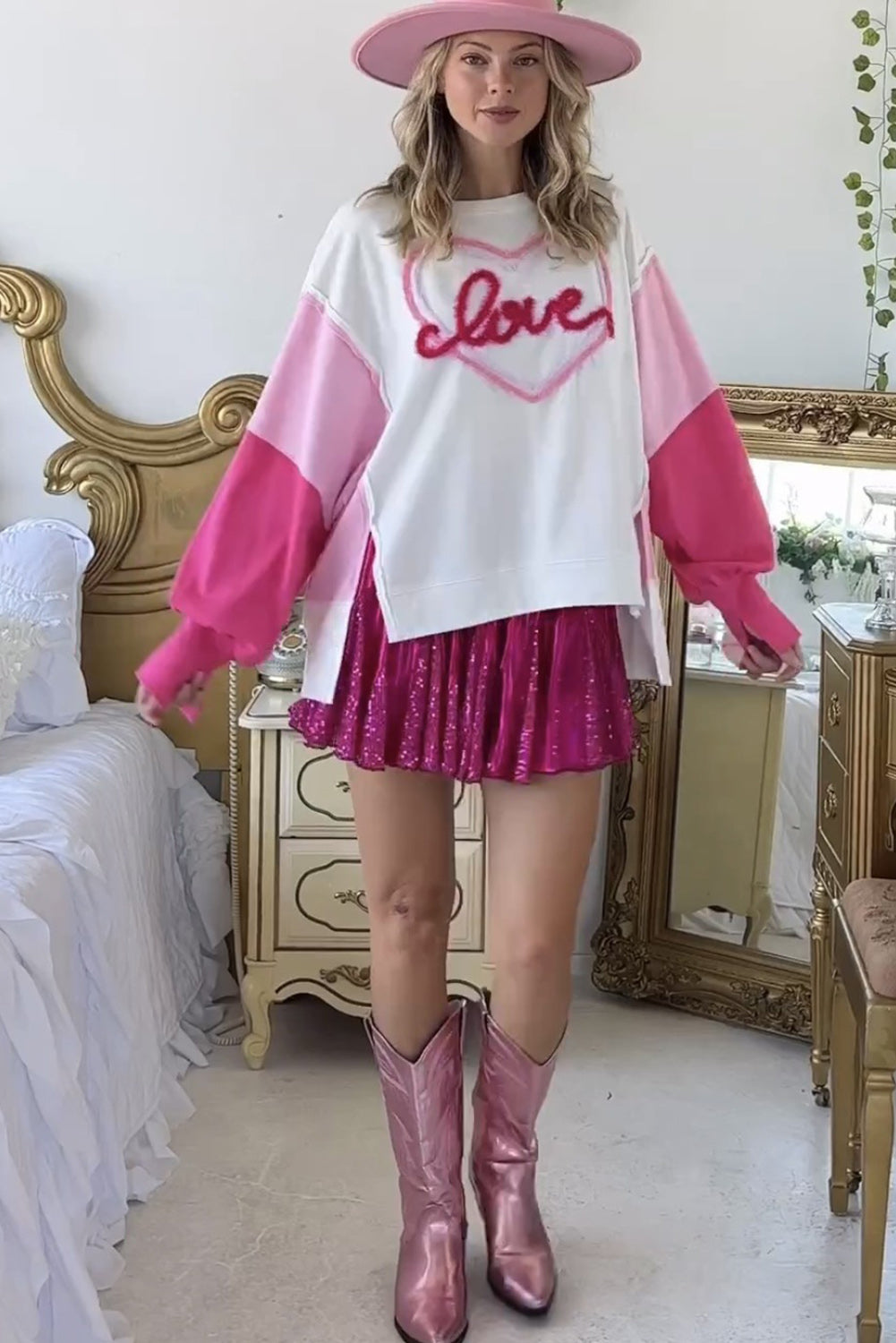 A woman with long blonde hair is smiling and posing indoors. She is wearing a White Tinsel Love Heart Graphic Color Block Sleeve Sweatshirt with pink shorts. There are framed photos and a table in the background.