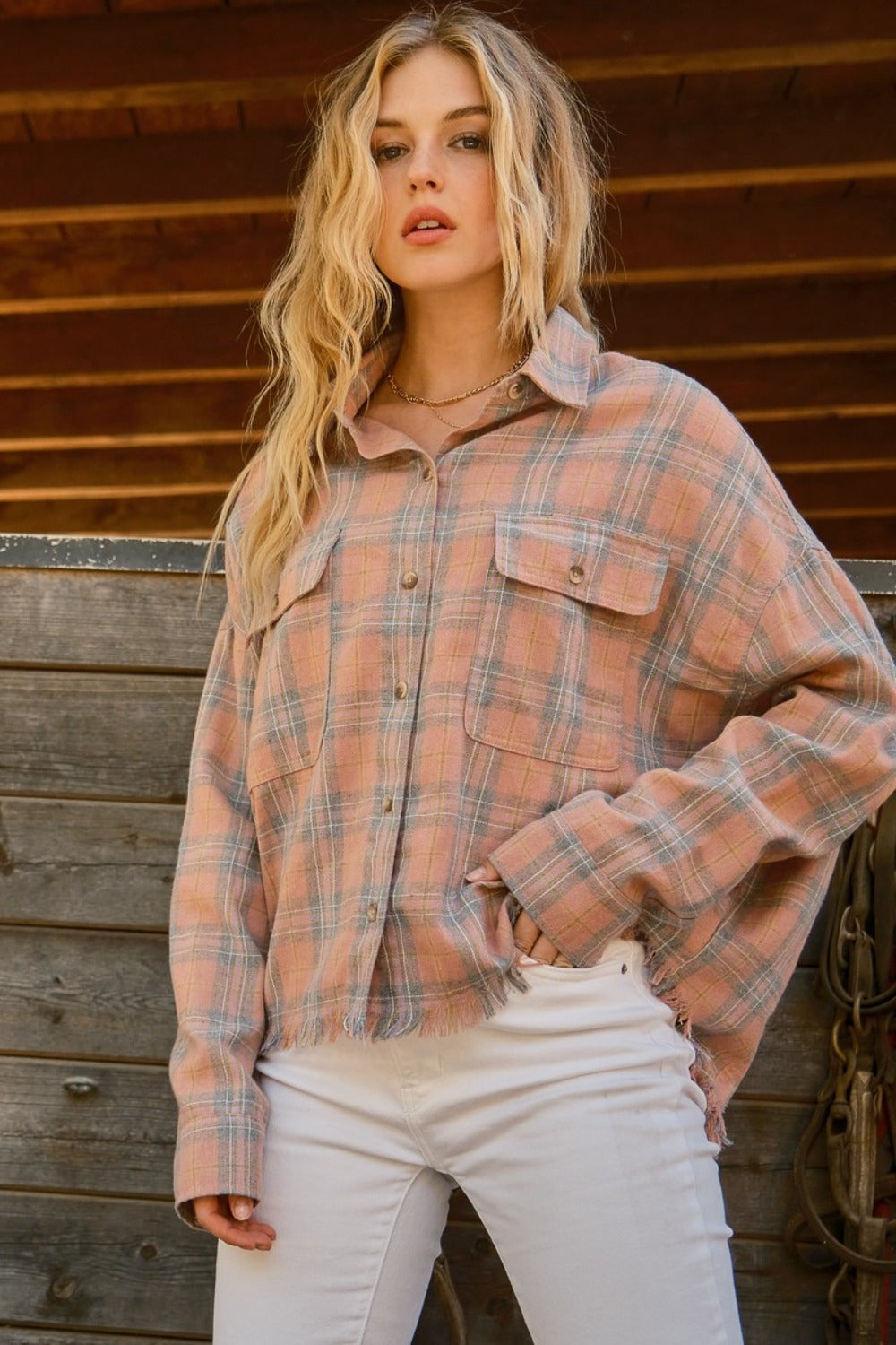 A person wearing the And The Why Full Size Plaid Button Up Raw Hem Shirt and white pants is standing in front of a wooden structure, showcasing raw edge detailing, with bridles hanging beside them.