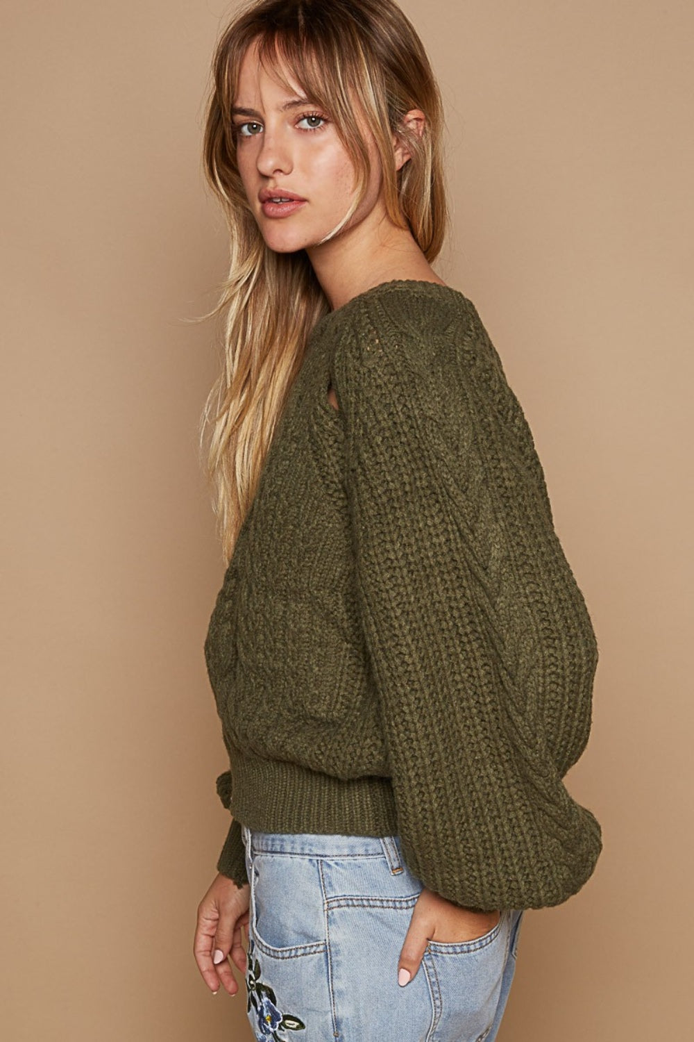 A woman wears the POL Cable Knit Cutout Long Sleeve Sweater in olive-green, showcasing a subtle shoulder keyhole, beautifully matched with light blue jeans adorned with floral embroidery, all set against a beige background.