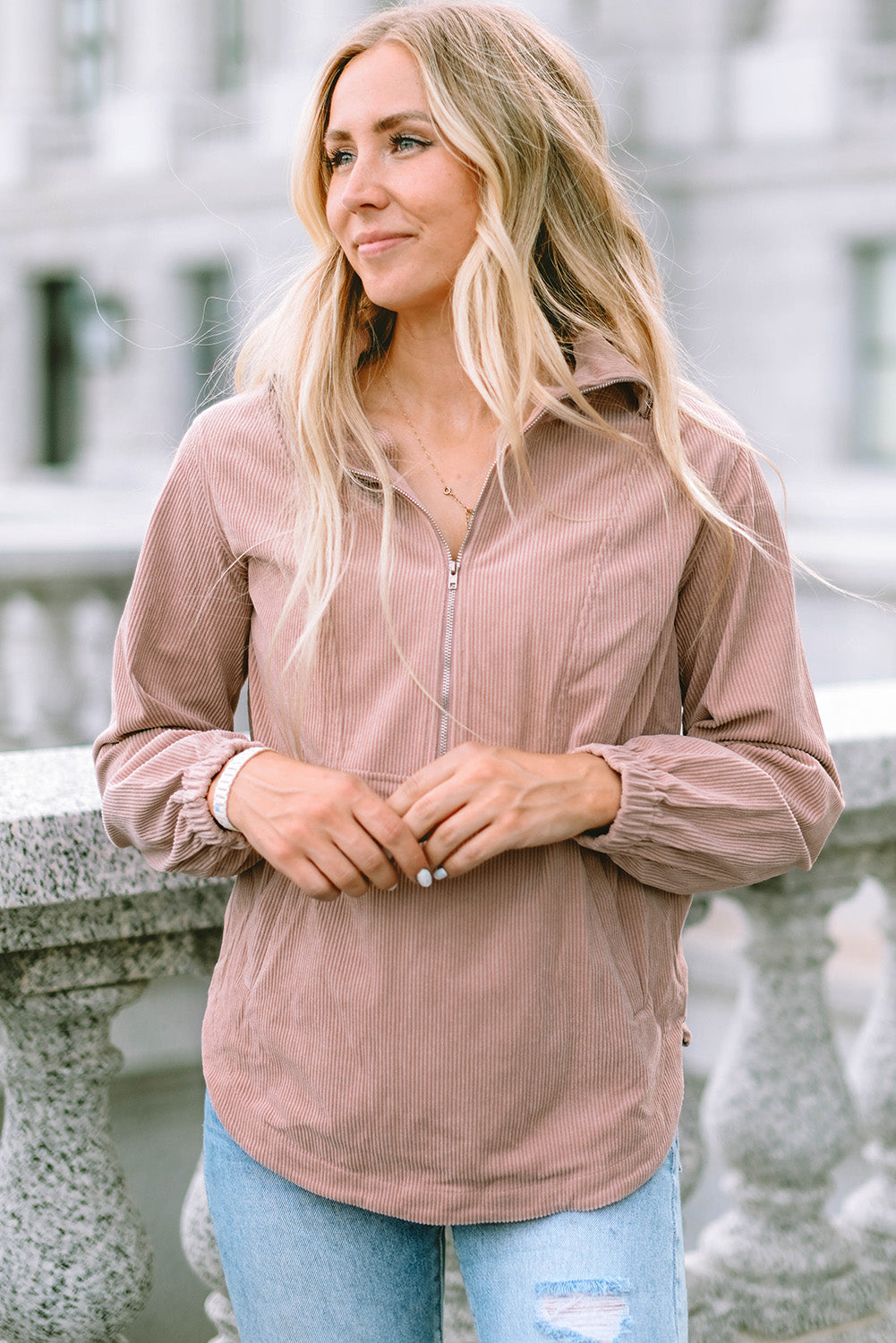 A person with long blonde hair, wearing a trendy Apricot Ribbed Texture Half Zip Collared Sweatshirt and blue jeans, stands facing away and looks over a stone balustrade.