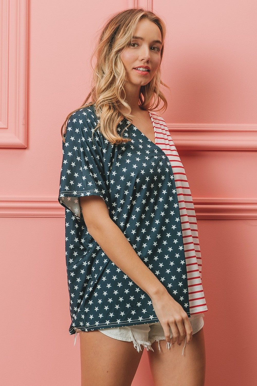 A woman stands against a pink wall, wearing the BiBi US Flag Themed Color Block Short Sleeve T-Shirt, which features one half blue with white stars and the other half red and white stripes. She is smiling with long wavy hair, showcasing her American pride casual wear.