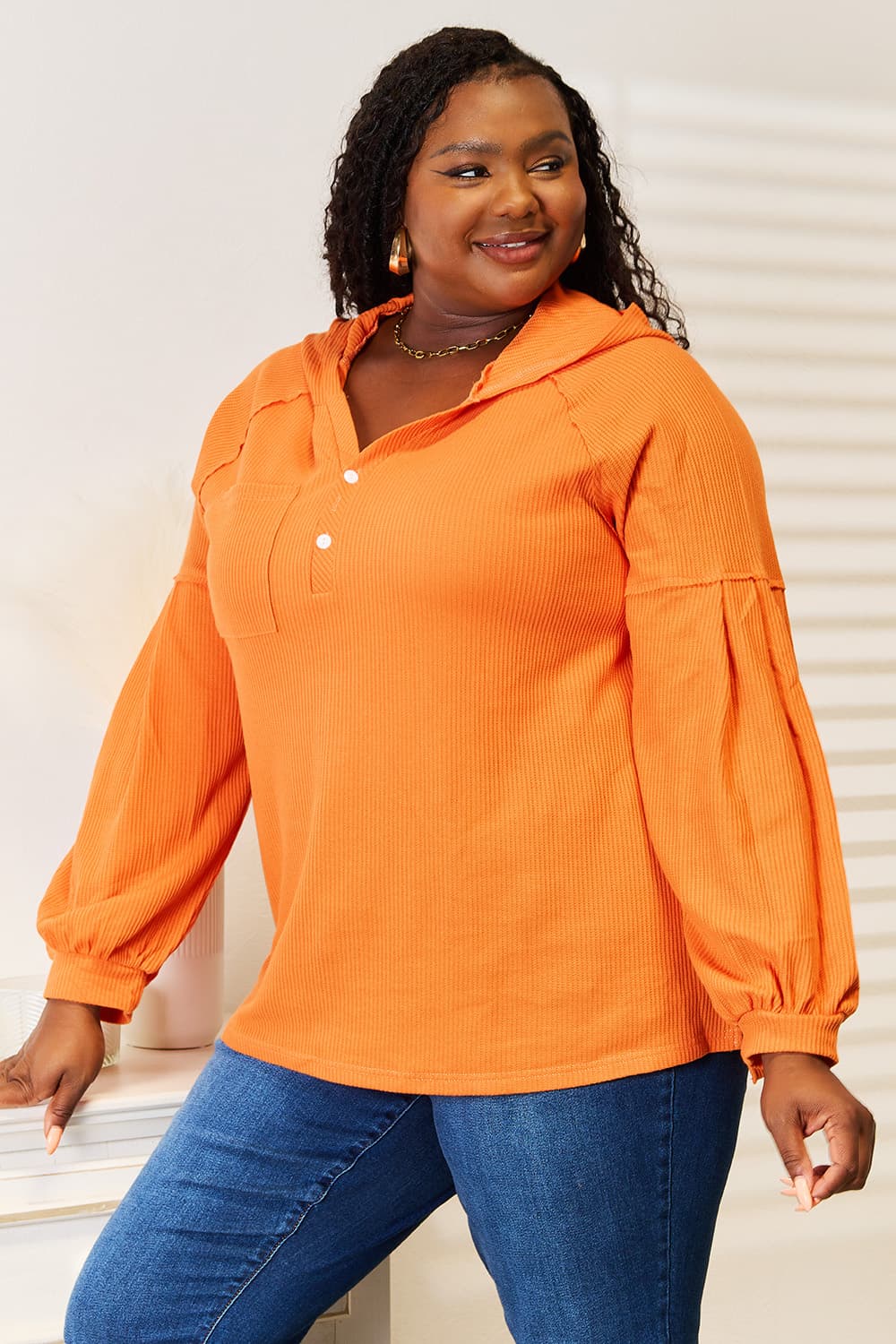 A person wearing an orange Basic Bae Side Slit Buttoned Waffle Knit Hoodie with blue jeans stands indoors near a white dresser, smiling and looking to the side.