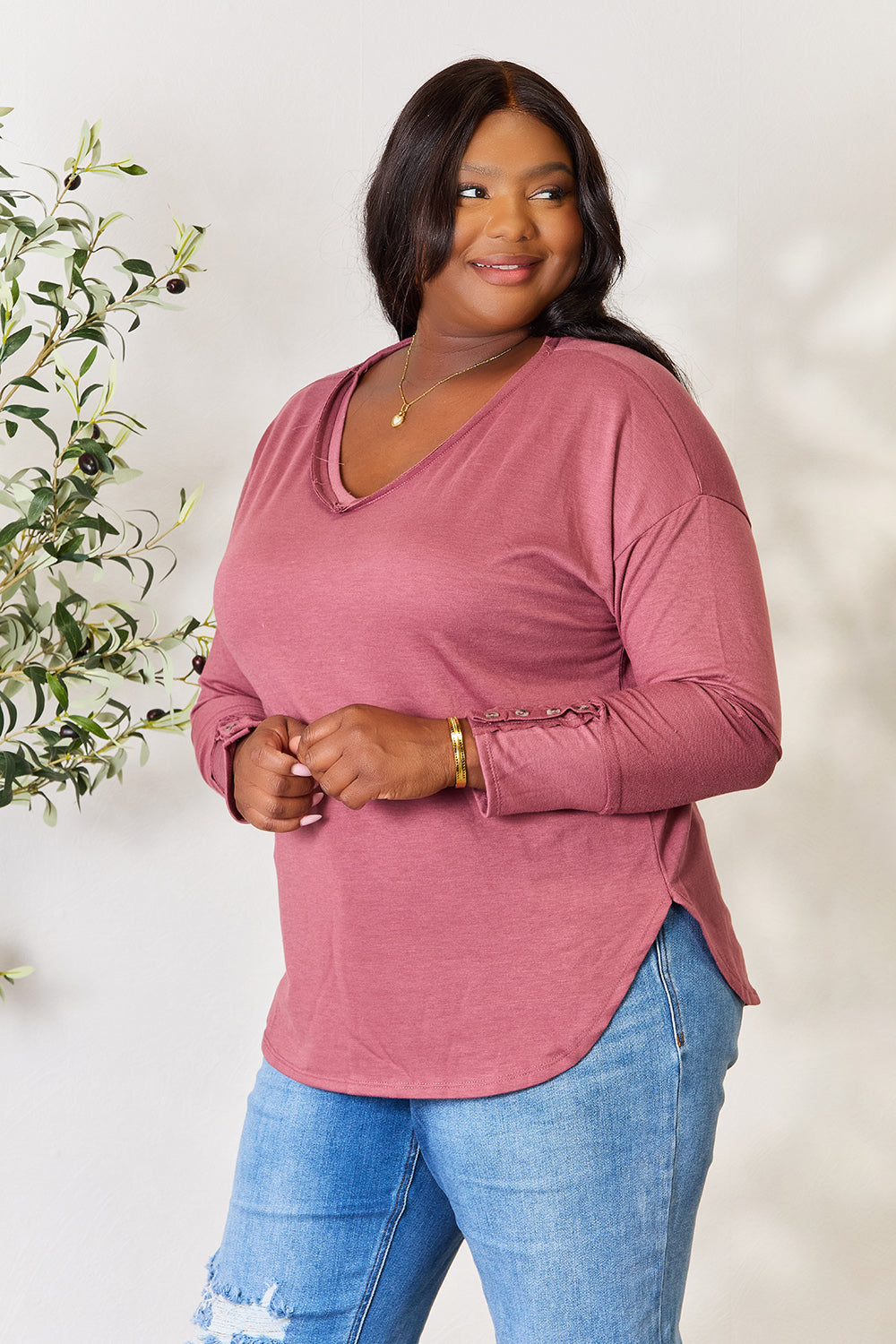 A person stands in front of a plain backdrop with a plant, wearing a maroon Culture Code Full Size V-Neck Exposed Seam Long Sleeve Blouse and blue jeans, smiling slightly with their hand on their hip. The relaxed fit of the polyester blend adds to the casual yet stylish look.