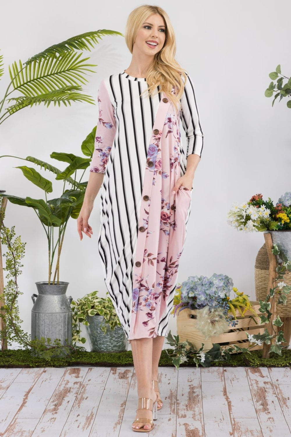 A woman stands amidst potted plants and flowers on a wooden floor, wearing the elegant and chic Celeste Full Size Floral Striped Contrast Midi-Dress with Pockets.