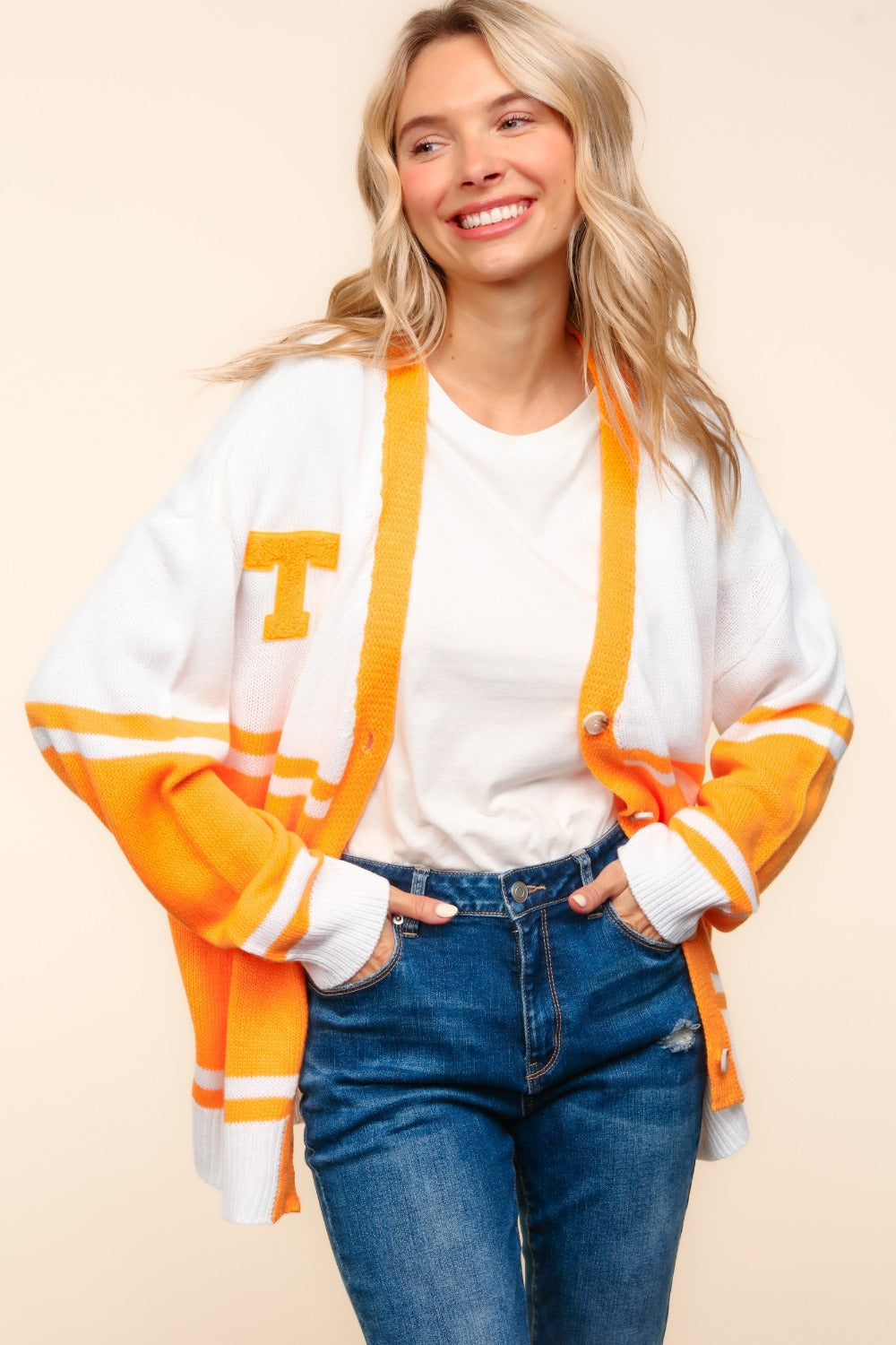 A person wearing the Haptics V Neck Button Down Letter Patch Cardigan, oversized with white and orange stripes and a yellow "T" patch, paired with blue jeans, smiles while lifting their hands to their head.