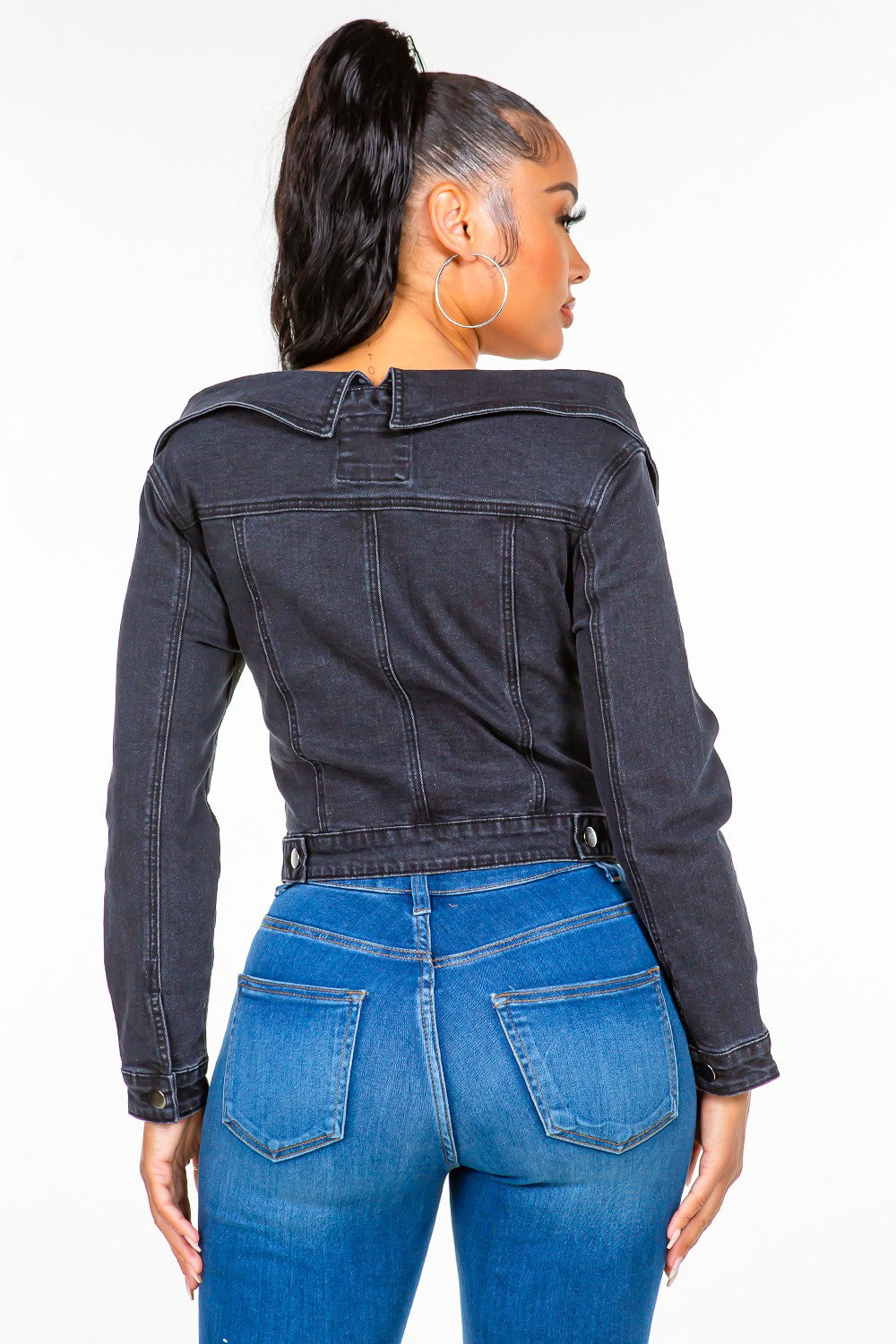 A woman in an American Bazi Off Shoulder Lace Up Denim Jacket and blue jeans stands against a plain white background. She has long hair tied up in a high ponytail.