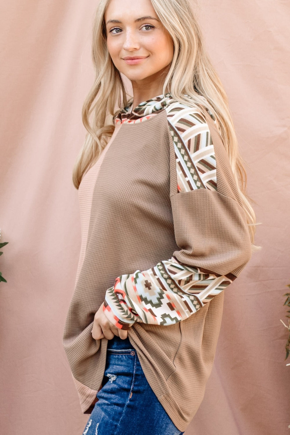 A woman with long blonde hair wears an And The Why Printed Color Block Hooded Waffle Top in various earth tones, standing in front of a light brown background.