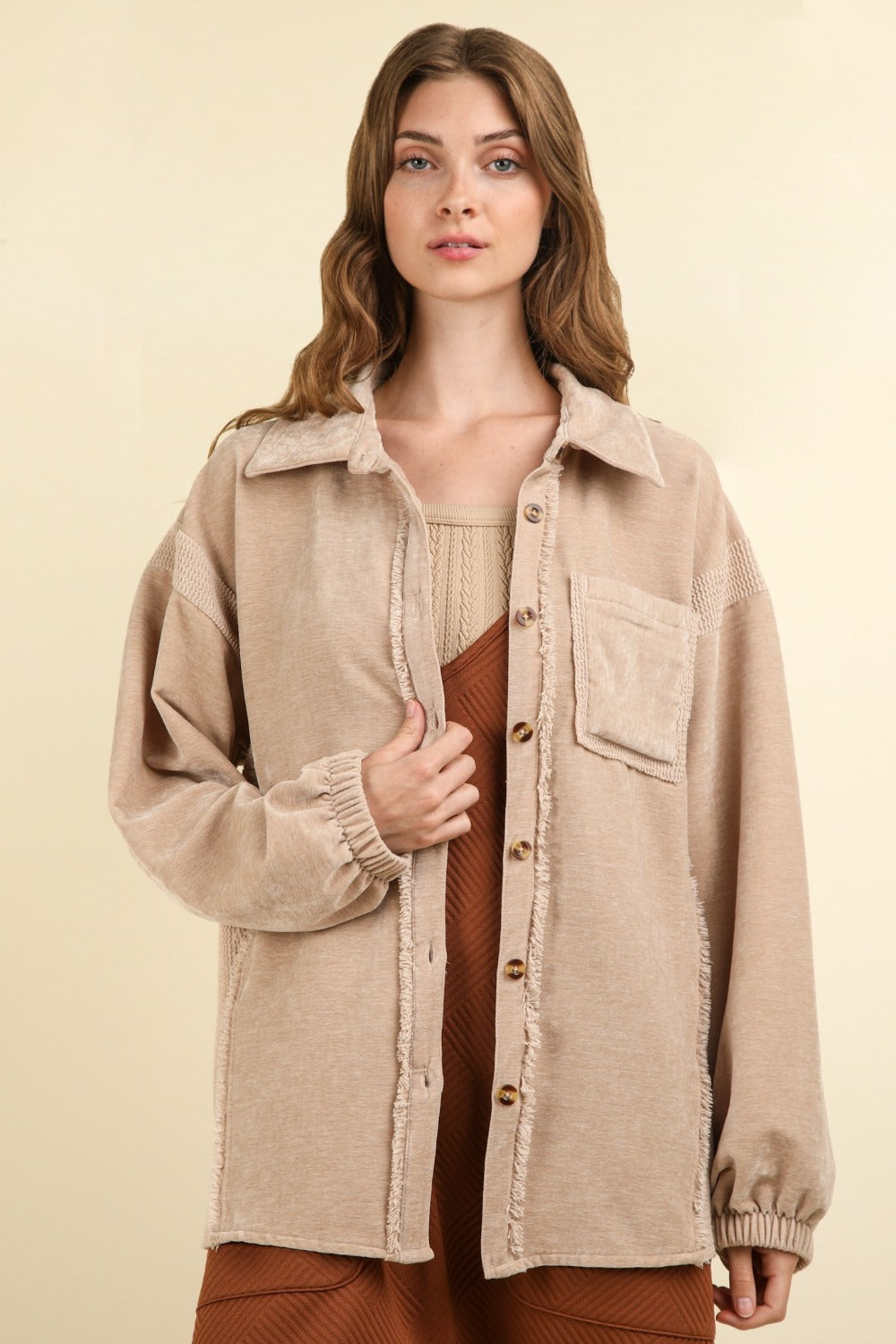 A woman with long wavy hair wears the VERY J Mixed Media Button Down Raw Hem Shacket featuring a drop shoulder design over a brown outfit. She faces the camera with one hand holding the lapel of her shacket against a plain beige background.