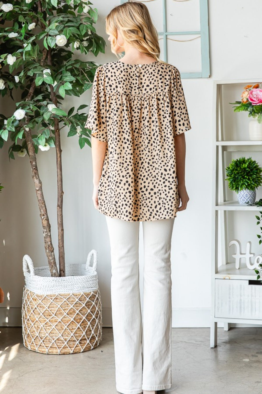 Wearing the Heimish Full Size Animal Print Flutter Sleeve Blouse, a person stands indoors near a plant and framed wall decor, stylishly pairing it with white pants.