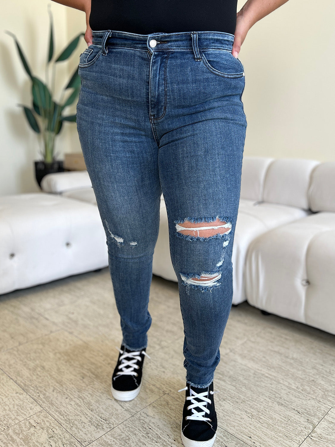 A person wearing Judy Blue Full Size High Waist Distressed Skinny Jeans and black sneakers stands indoors on a wooden floor, with a white sofa and green plant in the background.