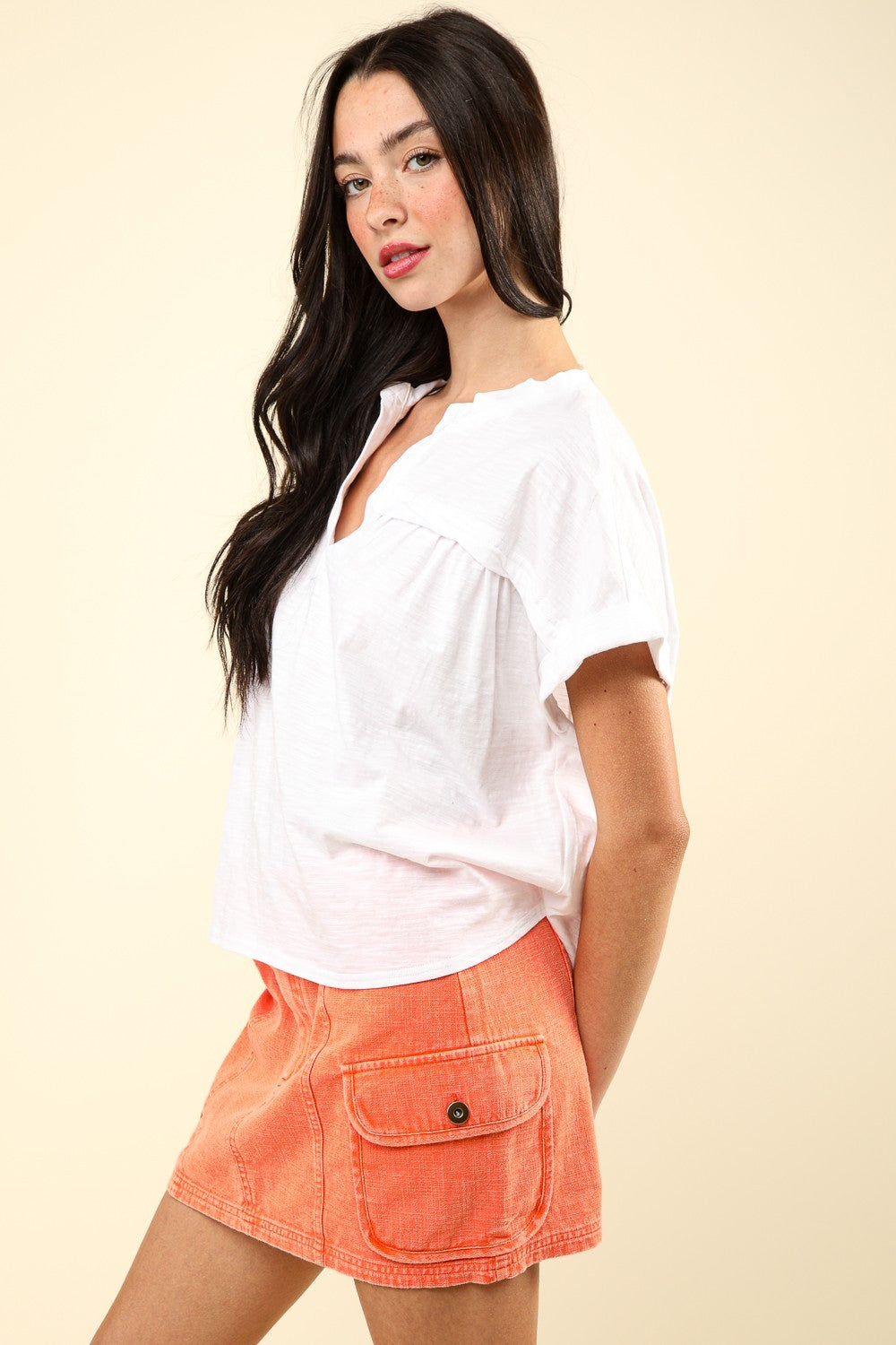 A woman wearing a white, VERY J Notched Short Sleeve Washed T-Shirt paired with an orange skirt poses against a neutral background, showcasing a versatile wardrobe staple.