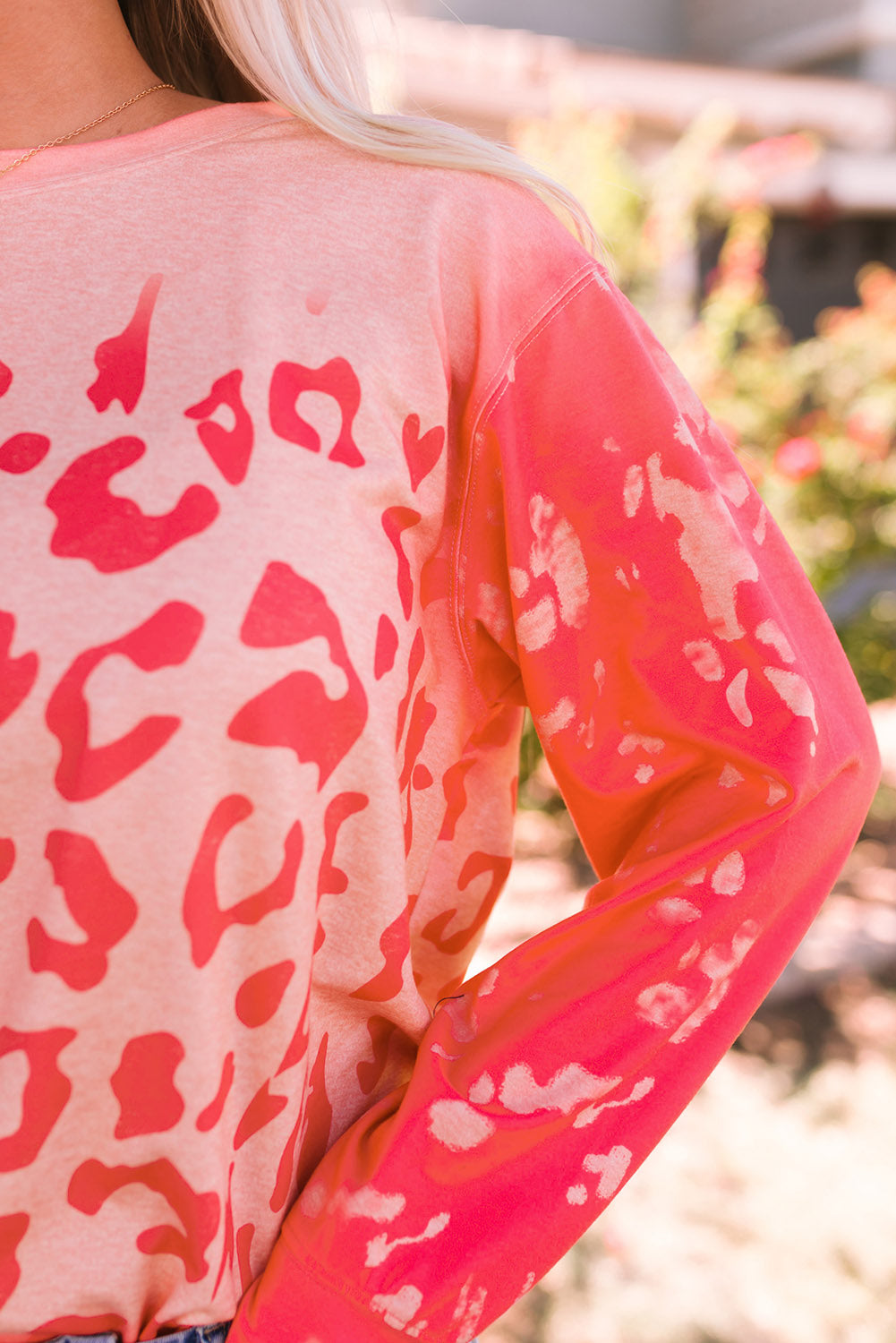 A woman with long blonde hair stands outdoors wearing the Pink Bleached Cheetah Print Sweatshirt and blue jeans. She is smiling and posing with one hand in her pocket.