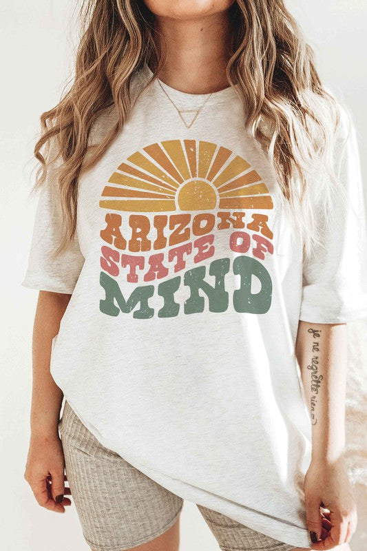 Person wearing the ARIZONA STATE OF MIND Graphic Tee, a beige 100% cotton t-shirt with "ARIZONA STATE OF MIND" text in brown and green colors. The unisex shirt offers both comfort and style for any occasion. They are holding a pink polka-dotted mug.