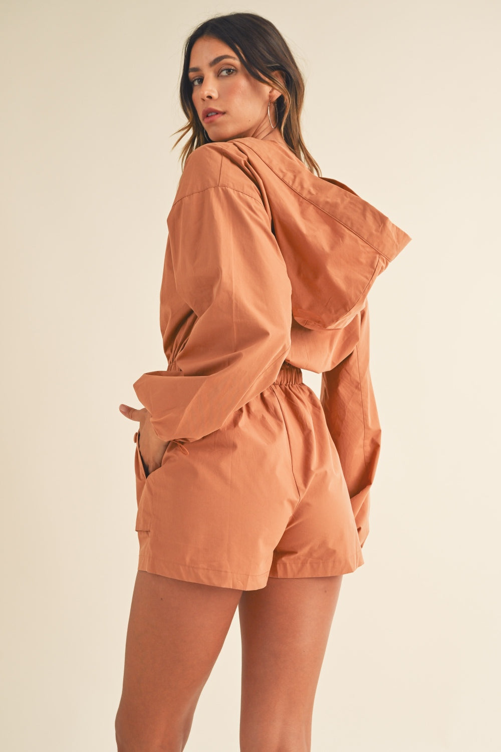 A person wearing the Mable Snap Down Long Sleeve Hooded Romper, featuring a brown color, long sleeves, a snap-down front, and an elastic waistband, is standing against a plain background.