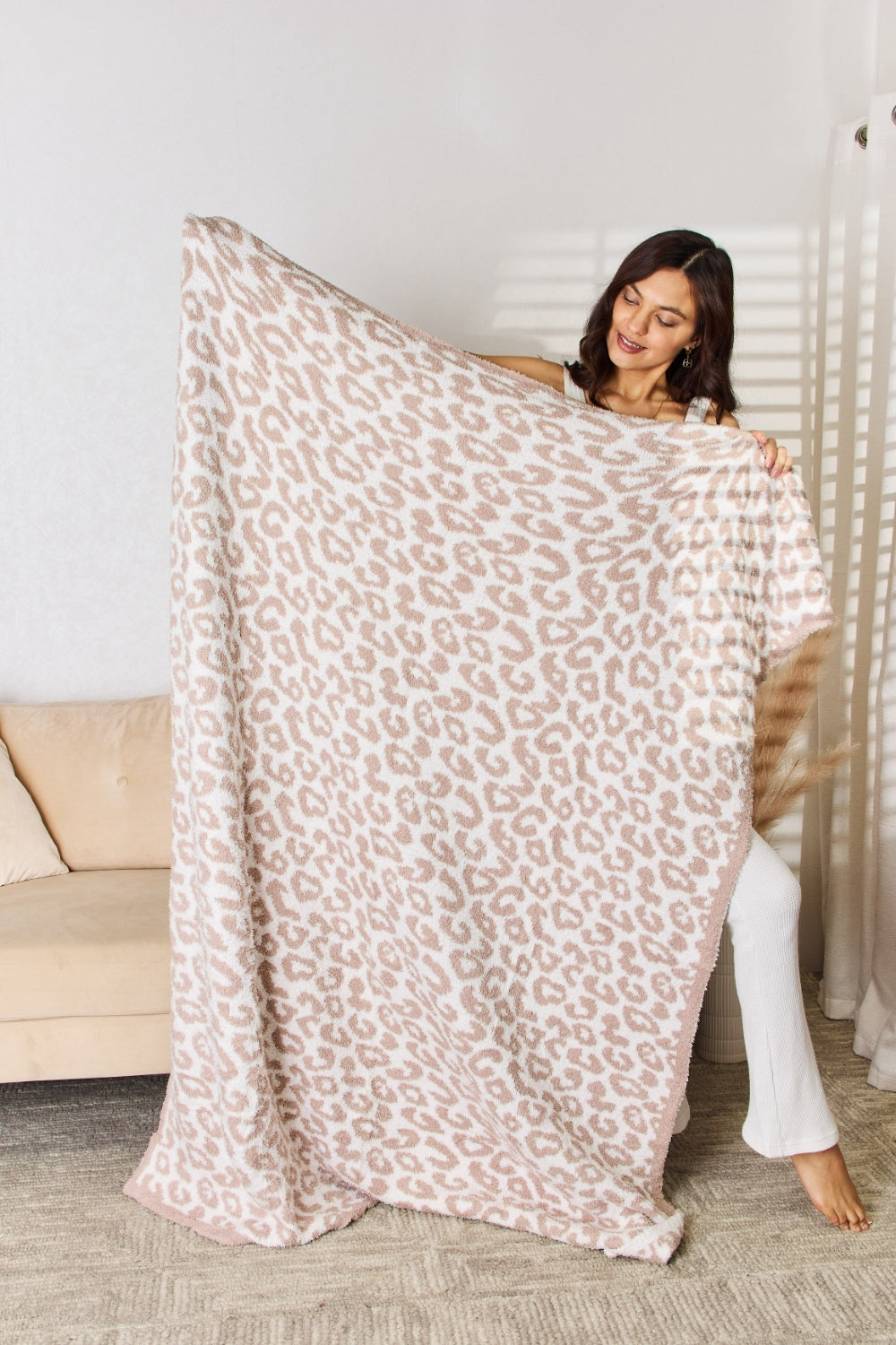 A woman stands in a room holding up the Cuddley Leopard Decorative Throw Blanket, which features a large, leopard-print design in a neutral color scheme. She is smiling and facing the camera, showcasing the 100% polyester fabric.