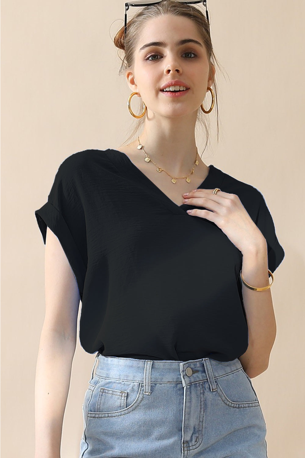 A woman wearing a stylish Ninexis V-Neck Trim Rolled Short Sleeve Shirt in light blue and high-waisted blue jeans, accessorized with hoop earrings, a necklace, and sunglasses on her head, poses in front of a beige background.