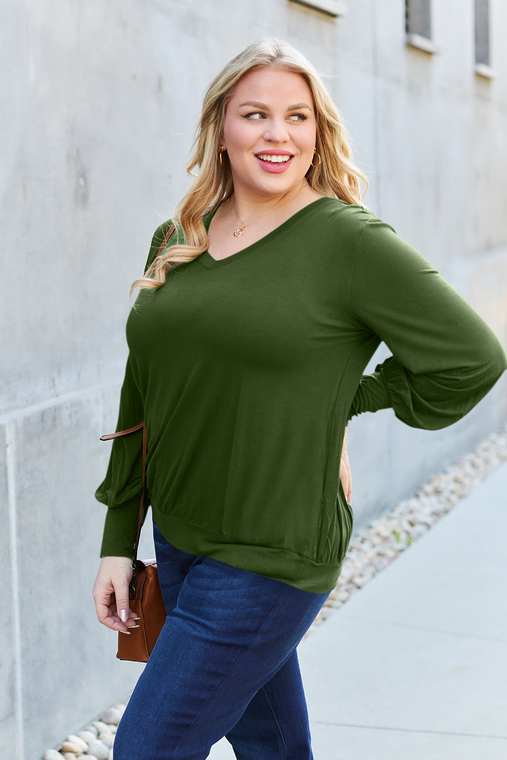 A woman with brown hair is standing against a concrete wall, wearing the Basic Bae Full Size V-Neck Lantern Sleeve Top in teal and blue basic-style jeans, smiling.