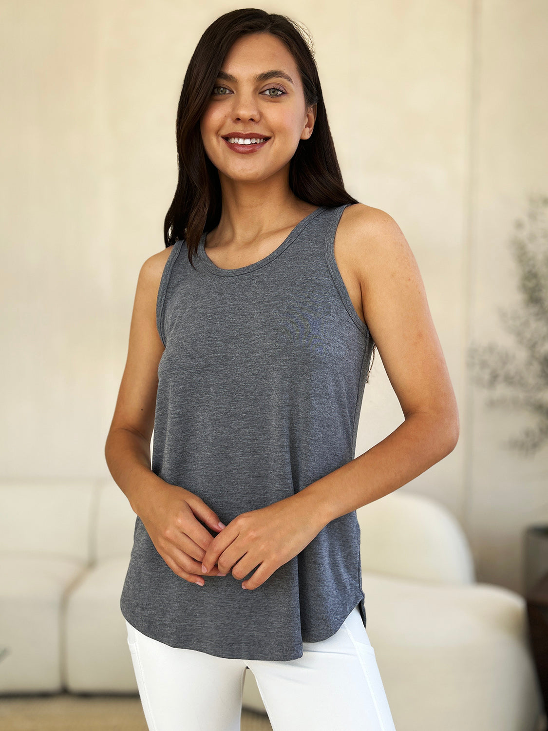 A woman wearing sunglasses, blue jeans, and the Basic Bae Full Size Round Neck Curved Hem Tank is standing in front of a concrete wall, holding her glasses with one hand and carrying a beige shoulder bag. Her basic style complements the slightly stretchy fabric of her outfit, ensuring comfort while maintaining chic simplicity.