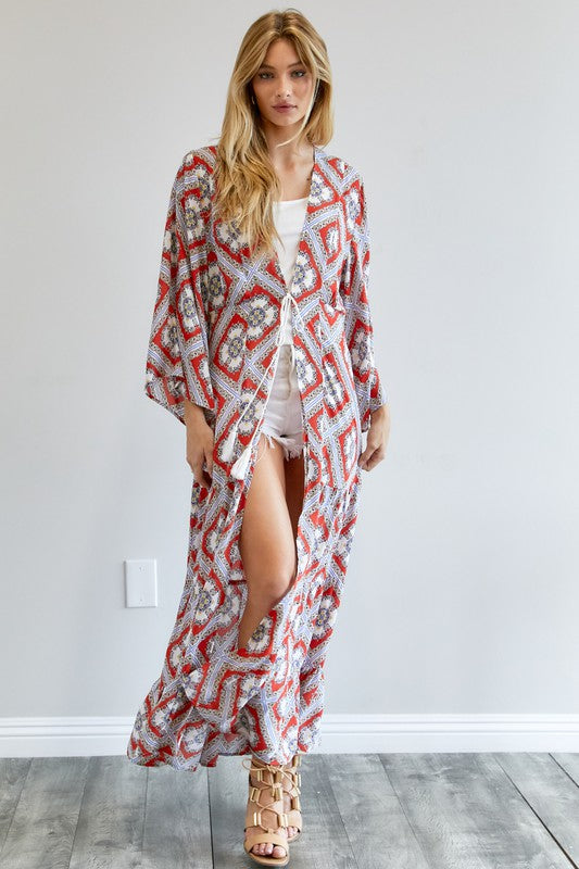 A person wearing a Printed Long Sleeve Loose Kimono with a bohemian blue and pink print over a white outfit stands in a room with a gray floor and white wall.