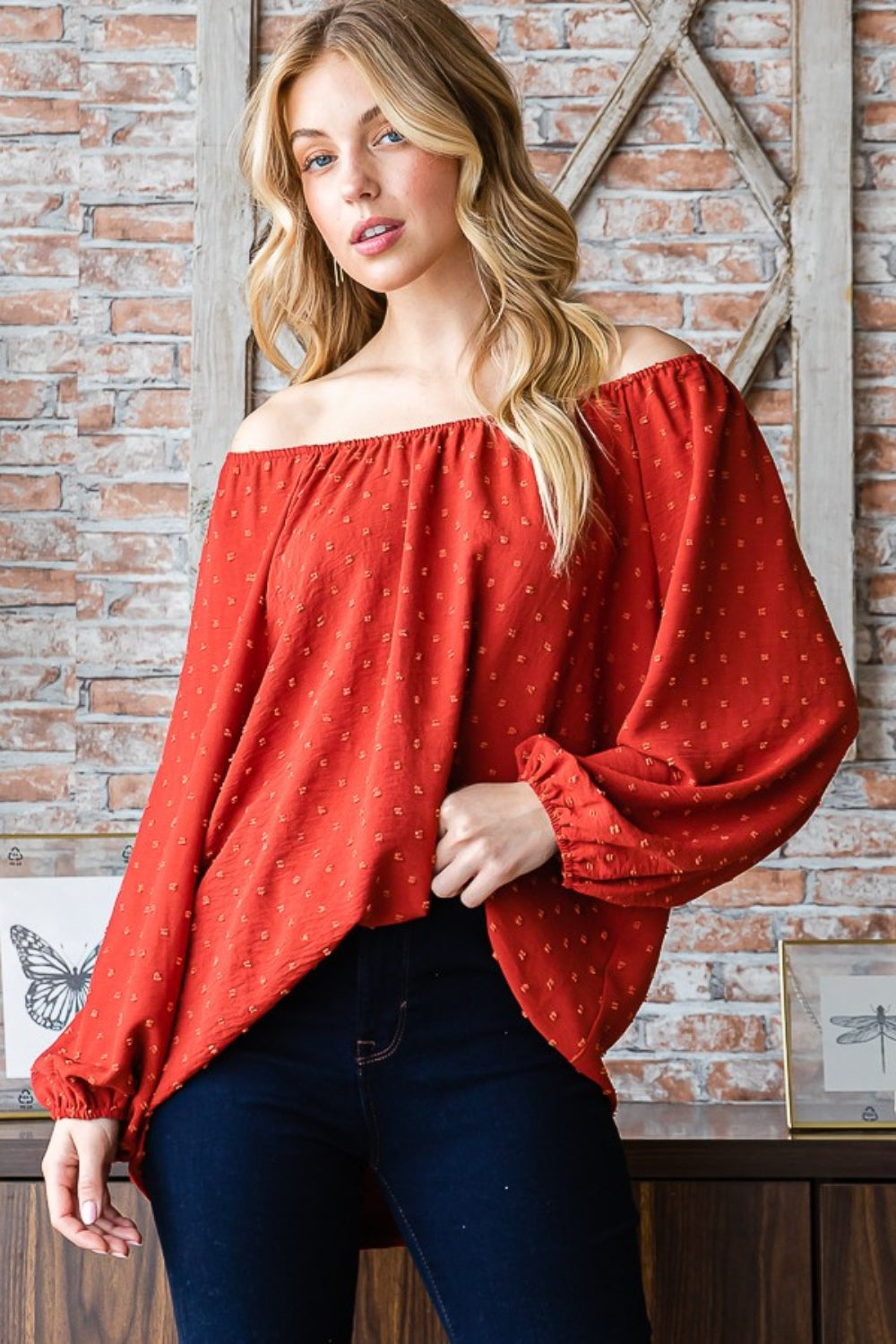 A woman wearing a Heimish Full Size Swiss Dot Off Shoulder Top paired with dark pants poses indoors against a brick wall decorated with butterfly artwork.