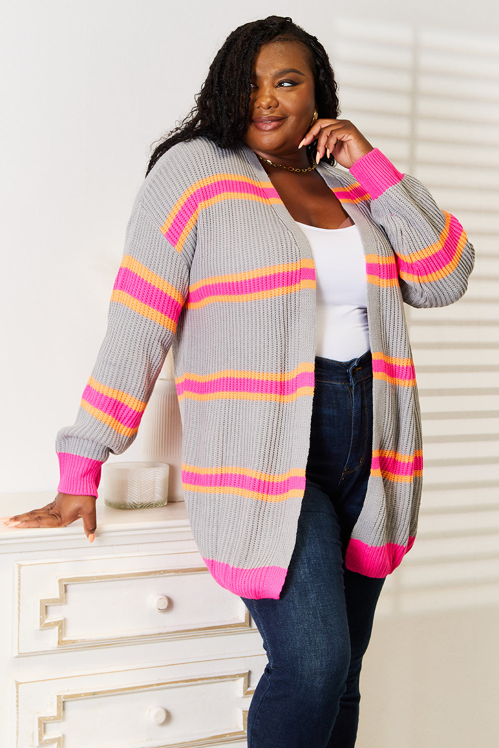 A woman stands against a white wall, smiling with one hand on her hip. She wears the Woven Right Ribbed Long Sleeve Cardigan, featuring pink and orange stripes, over a white top and dark jeans—an excellent choice for versatile layering.