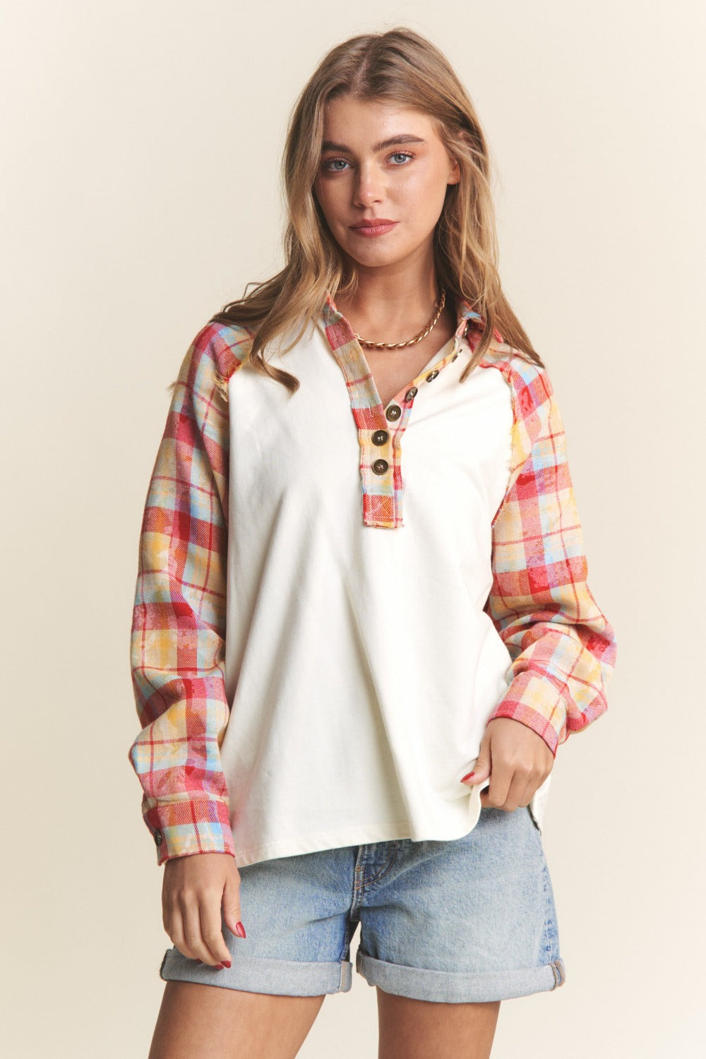 A woman wearing the J.NNA Plaid Contrast Button Down Henley Top—a stylish white pullover shirt with red and yellow plaid sleeves—and denim shorts, stands against a plain background.