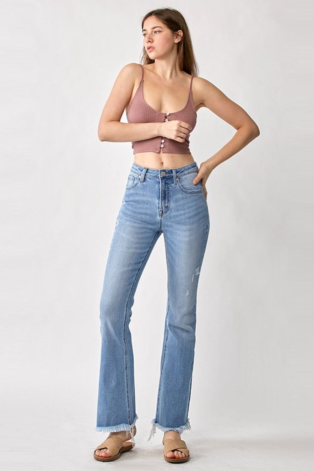 A person wearing RISEN High Rise Frayed Hem Bootcut Jeans in light-blue stretchy denim paired with tan sandals, standing against a plain background.