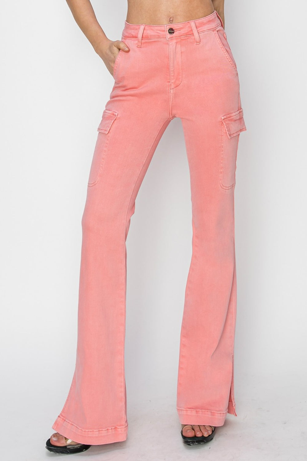 A person wearing RISEN Full Size High Rise Side Slit Cargo Bootcut Jeans, featuring a pink flared design with front pockets, stands against a plain background.