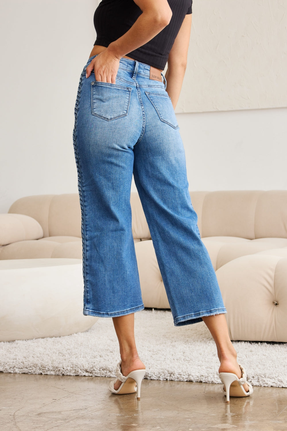 A person wearing Judy Blue Full Size Braid Side Detail Wide Leg Jeans and black heels is standing on a polished floor. There is a white couch and a large potted plant in the background.