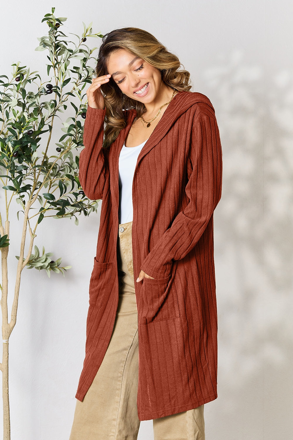 A woman wearing the Basic Bae Full Size Hooded Sweater Cardigan in black, paired with a white top and khaki pants, stands in front of a light background accented by a leafy plant on her left. With one hand tucked into her pocket, she gazes to her right.