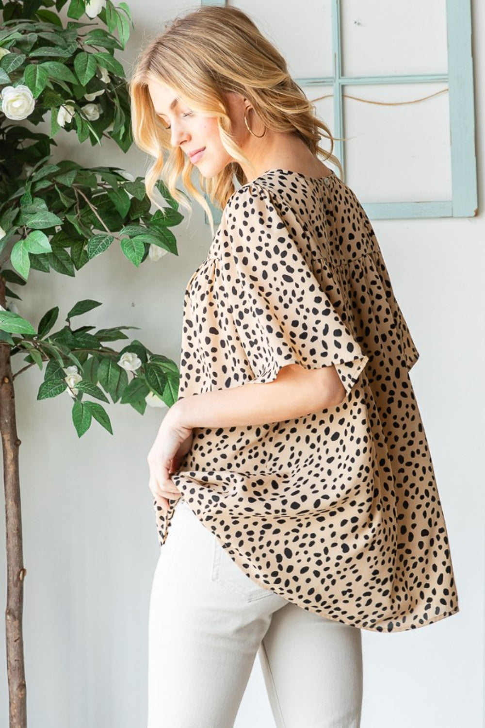 Wearing the Heimish Full Size Animal Print Flutter Sleeve Blouse, a person stands indoors near a plant and framed wall decor, stylishly pairing it with white pants.