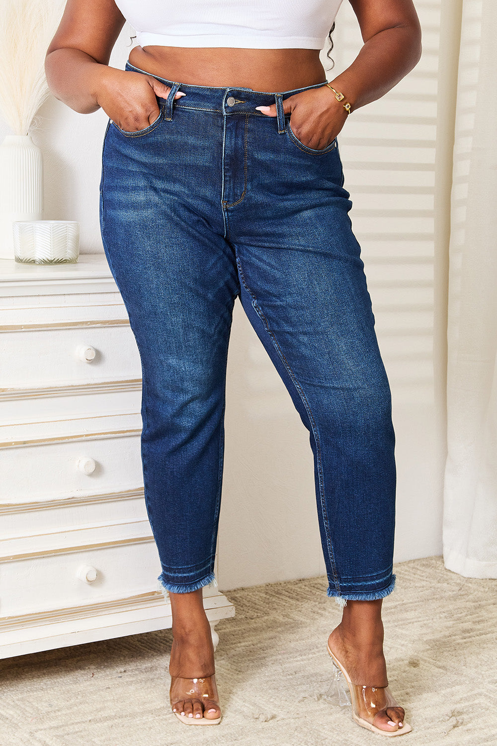 A person wearing Judy Blue Full Size High Waist Released Hem Slit Jeans in indigo wash and a white cropped top stands by a white dresser, showcasing a flattering fit.